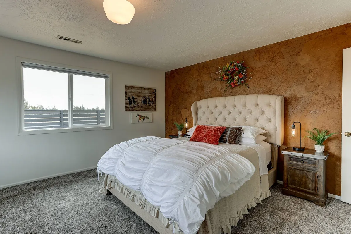 A bright bedroom with a plush tufted headboard, decorative wall finish, framed artwork, and a nightstand with a lamp. The space is filled with natural light and soft, calming tones, suggesting a restful ambiance.