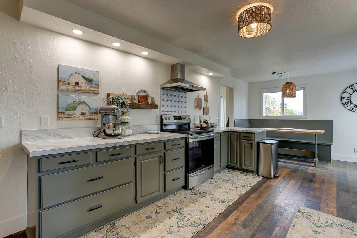 A warm, inviting kitchen with marble countertops, green cabinetry, stainless steel appliances, decorative wall art, and an open dining space. The room is filled with natural light, blending vintage character with modern updates.