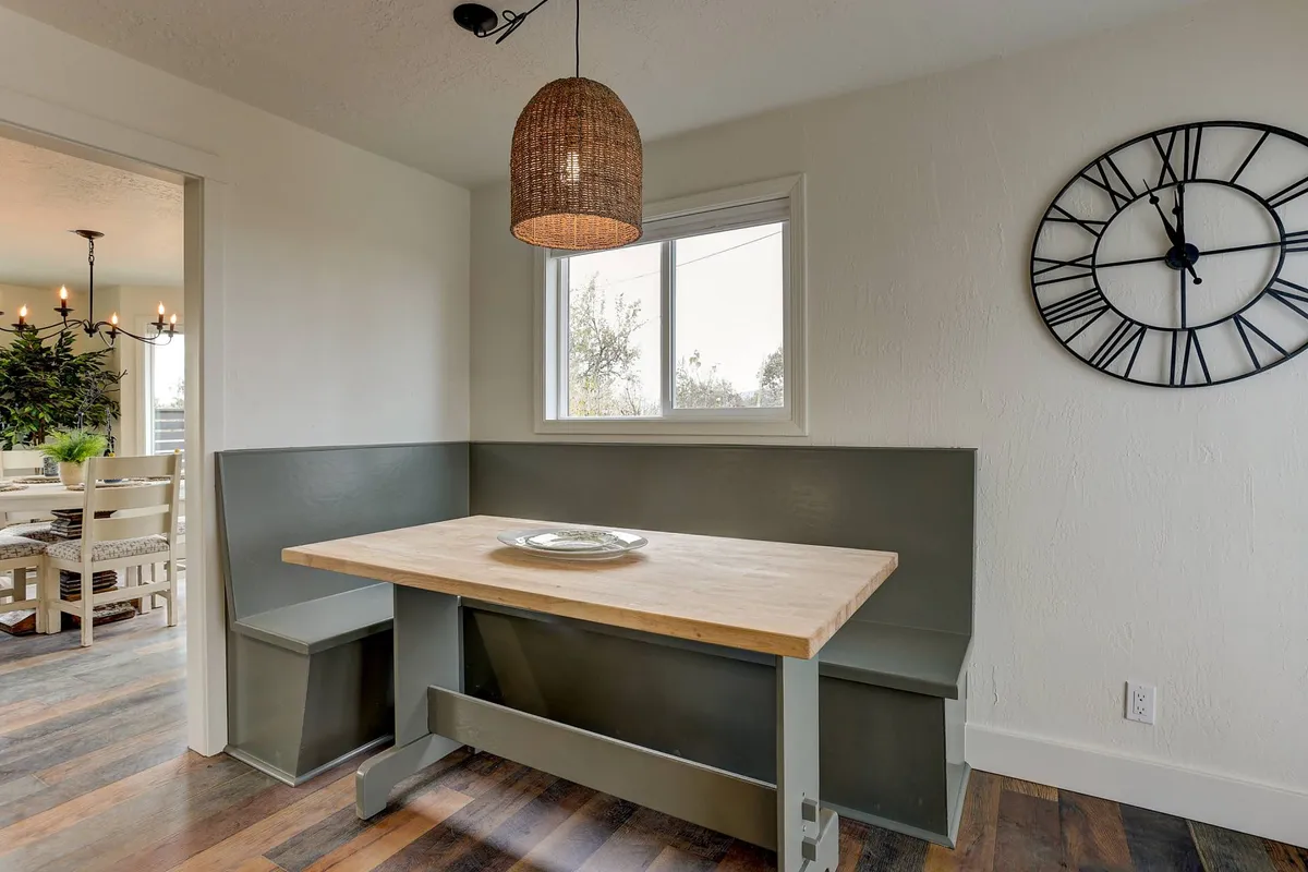A cozy interior dining space with a wooden table, woven pendant light, and a clock on the wall, designed to accommodate family meals or group gatherings. The setup suggests a comfortable setting for sharing stories, enjoying meals, and celebrating together.