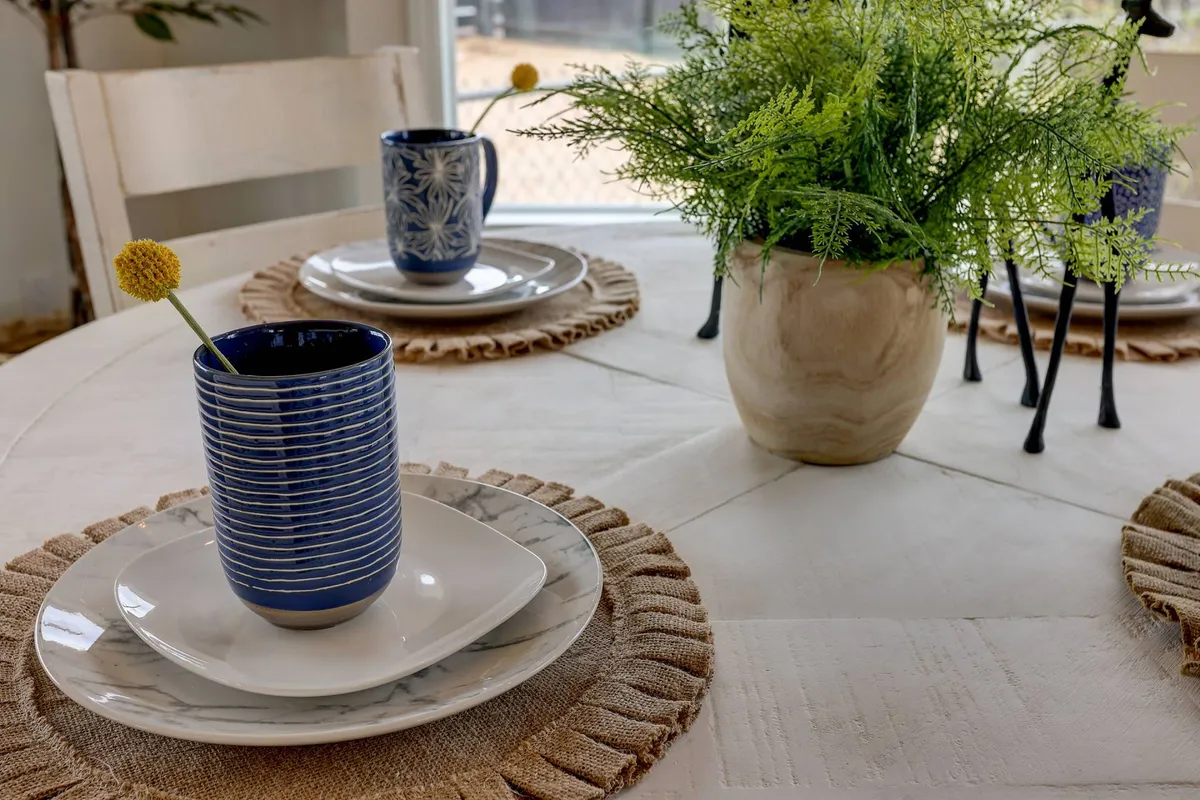 A warm, welcoming dining area with a blue striped mug, green plant, elegant plates, and textured placemats arranged on the table. The window view hints at natural surroundings, suggesting a connection between inside and out.