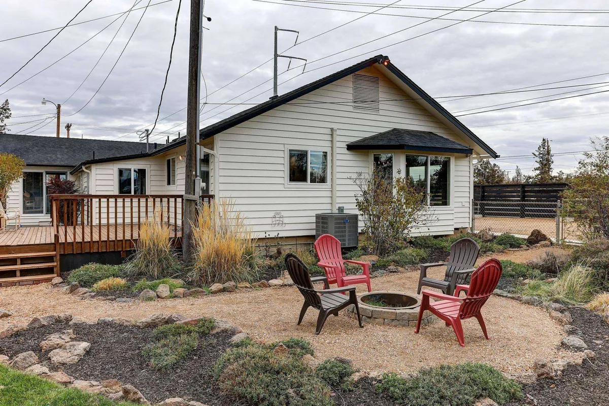 A cozy outdoor fire pit area with red and black chairs arranged in a circle, set within a landscaped garden. The fire pit is ready for an evening gathering, and the surrounding greenery provides a sense of privacy and intimacy. The setting feels ideal for stargazing, sharing stories, or enjoying a glass of wine together under the night sky.