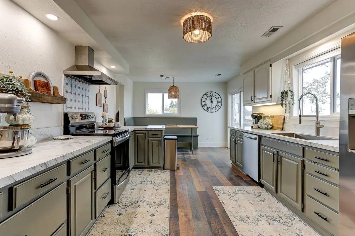 A warm, inviting gourmet kitchen featuring a central island, stainless steel appliances, natural wood accents, decorative pendant lighting, and a large window that lets sunlight pour in. The kitchen is open and connects seamlessly to other living spaces, hinting at an open layout ideal for gathering and cooking together.