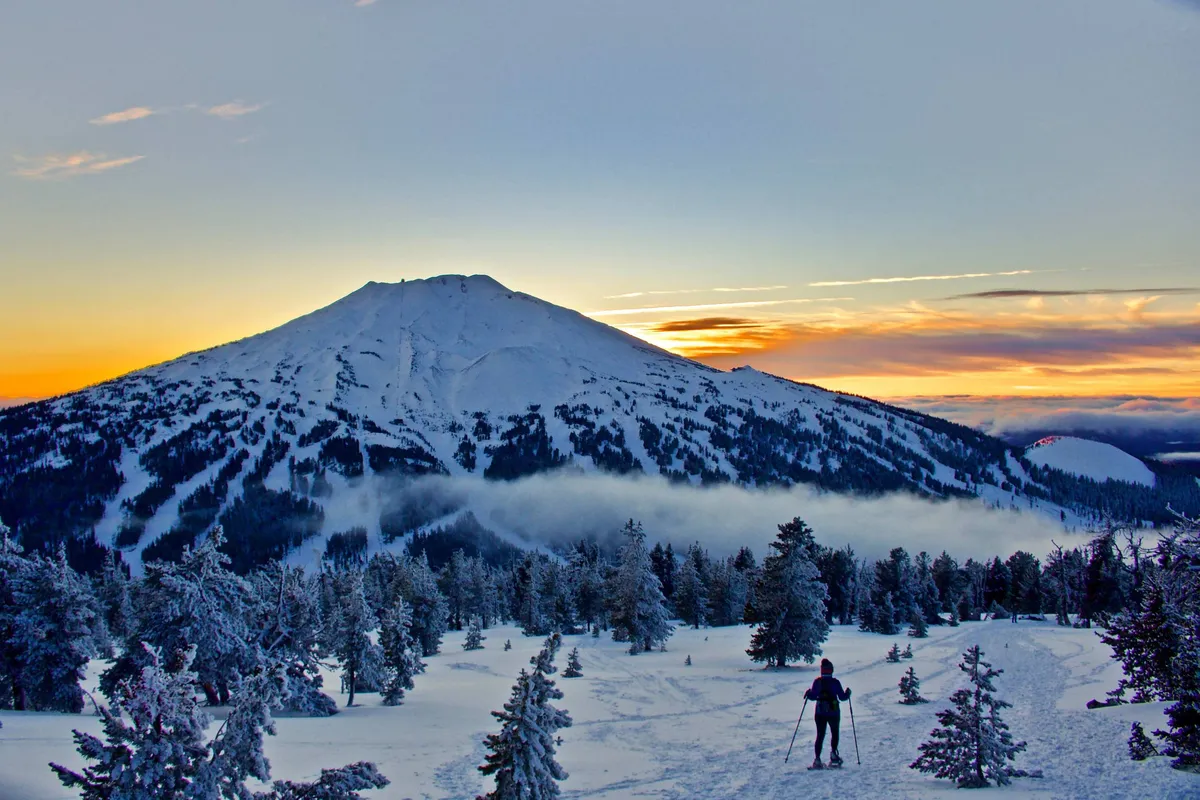 A stunning winter landscape with a snow-covered mountain, tall pine trees, a winding snowy path, and a colorful sunset sky. The scene is tranquil yet alive with the potential for winter sports and scenic exploration.