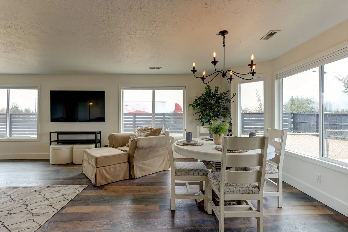 A spacious, sun-drenched dining and living area with a large table set for family meals, a statement chandelier, plush sofas, and expansive windows that let in natural light. The space exudes warmth, comfort, and a welcoming atmosphere, ideal for gatherings and celebrations.