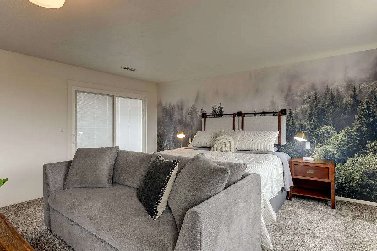 A tranquil bedroom featuring a comfortable bed adorned with decorative pillows and a cozy sofa, set against a striking mural of a serene forest. Gentle lighting and soft textures invite relaxation and quiet moments.