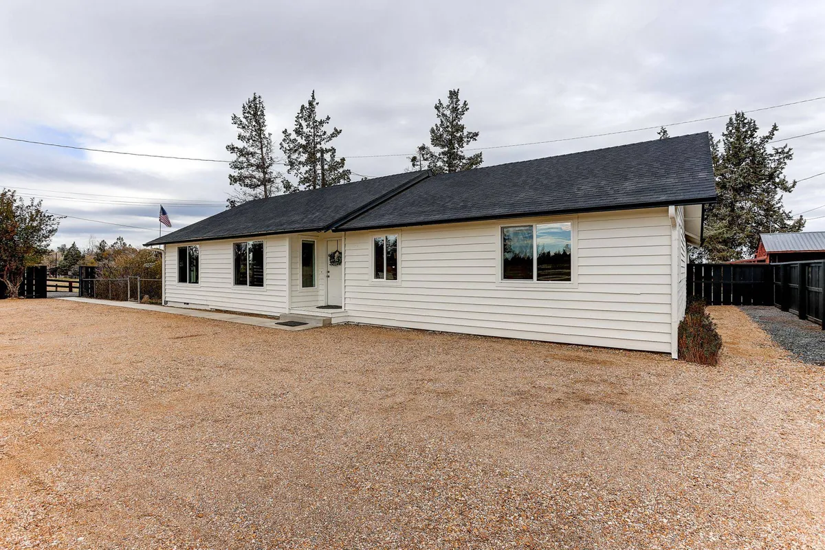A welcoming exterior view of the house featuring its facade, a spacious gravel driveway, and mature trees surrounding the property. The peaceful setting suggests seclusion and tranquility, offering a sense of privacy and natural beauty. The porch and yard invite guests to relax outside, perhaps with morning coffee or during sunset.